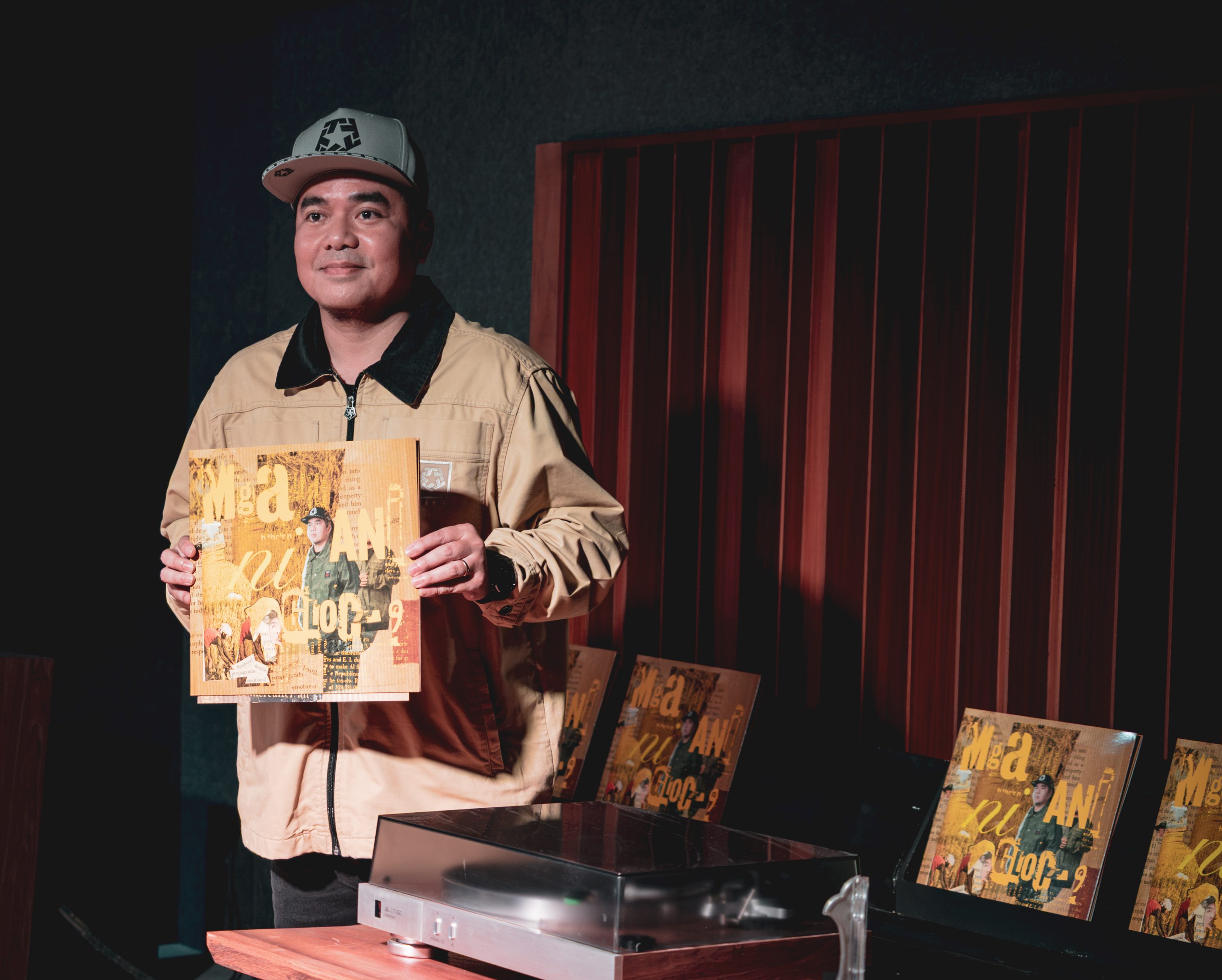 A man in a tan jacket holds up a copy of Mga Ani ni Gloc-9 beside a turntable, with more copies of the record on display behind him